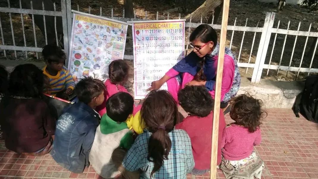 A volunteer teaching a group of young children from a Hindi alphabet chart and a numerical chart at an outdoor learning session